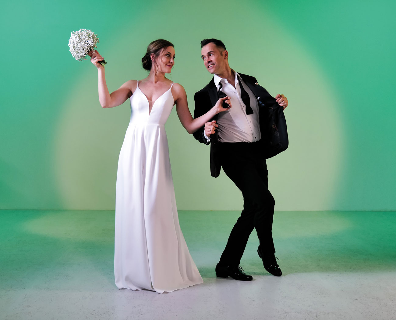 Jeune couple de mariés portant des chaussures de danse de Werner Kern Bridal devant un mur vert du studio.