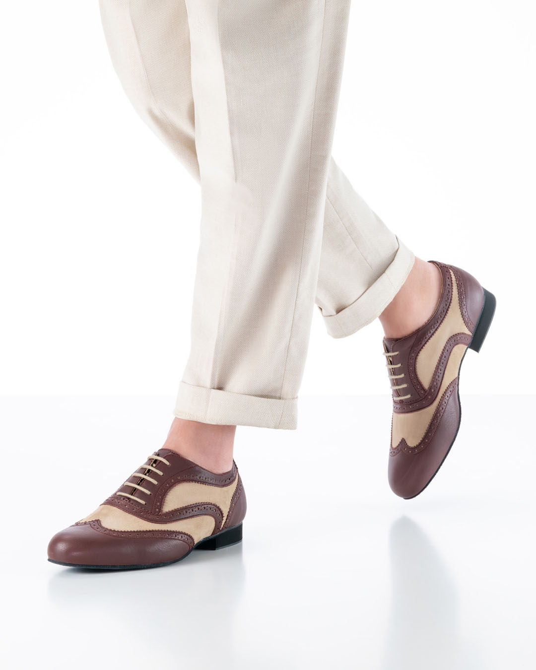 Un mannequin masculin, vêtu d'un pantalon couleur crème, s'avance vers la caméra avec des chaussures de danse couleur caramel et bordeaux, ornées d'un motif brogue ajouré.