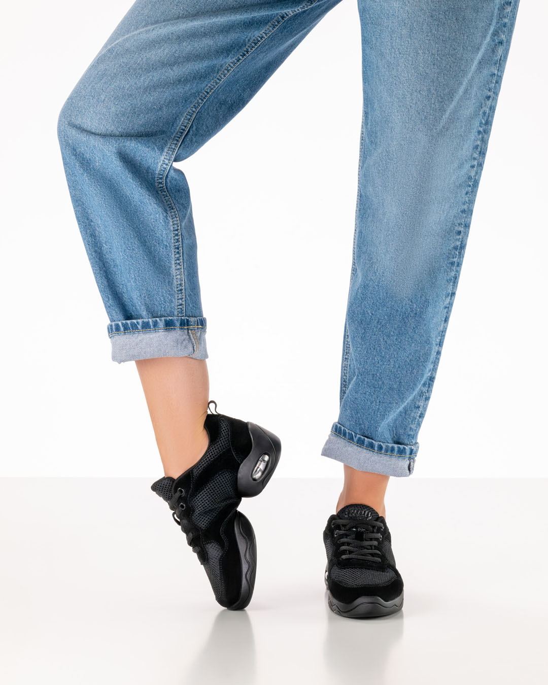 Un mannequin en jean porte des chaussures de danse noires à semelle fendue et plie le pied droit au niveau du cou-de-pied pour montrer la souplesse de la chaussure.