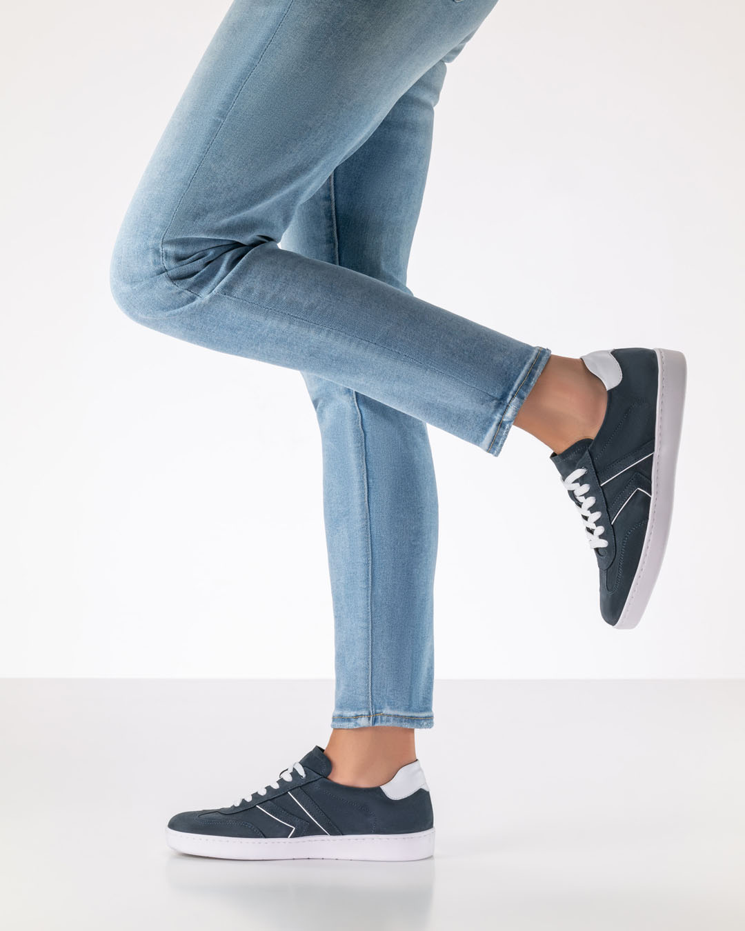Le mannequin se tient de profil et porte un jean clair et étroit ainsi que des baskets en daim bleu foncé à semelle blanche.